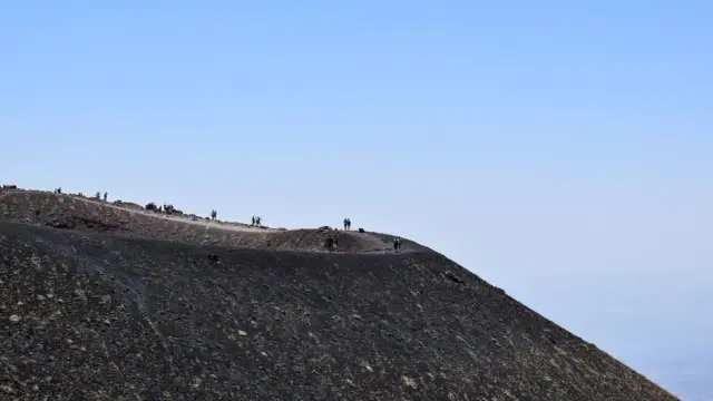 Hikers walking along the black volcanic crater rim of Mount Etna, Sicily, under a clear blue summer sky.