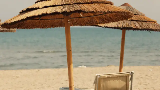 Traditional straw beach umbrellas and sun loungers on a golden-sand beach in Catania, Sicily, with the calm sea visible behind them.