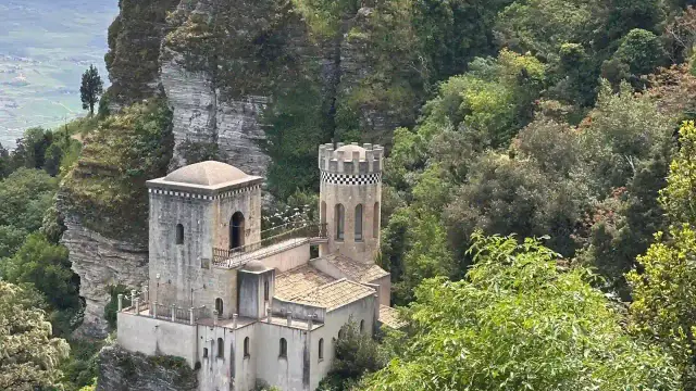 The Torretta Pepoli and the medieval walls of Castello di Venere perched on a cliff edge above Erice, Sicily, surrounded by dense green woodland.