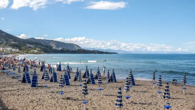 A long gold-sand beach in Giardini Naxos, Sicily, lined with rows of striped lido umbrellas and sunbeds, with a headland curving into the sea in the distance.