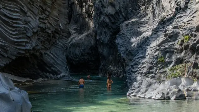 Tall black basalt rock columns rising on both sides of the Alcantara Gorge in Sicily, with a turquoise river running through the narrow canyon floor.
