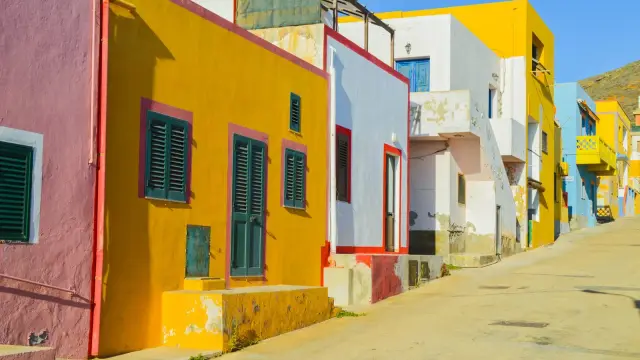 A street in Linosa's village lined with cube-shaped houses painted in bright yellow, pink, blue, and white, under a clear blue sky.