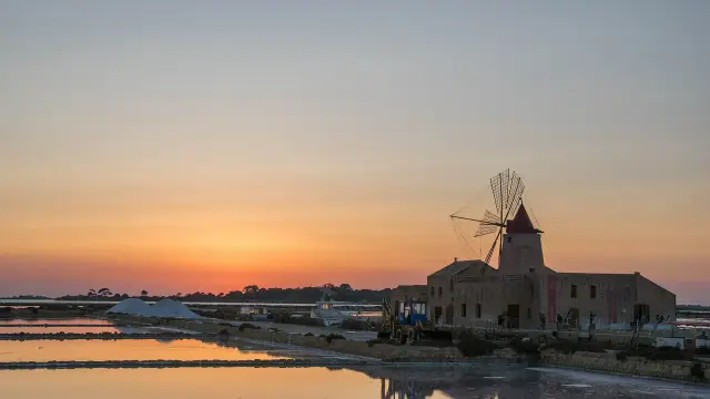A restored stone windmill and salt-worker's building at the Saline Ettore Infersa in the Stagnone lagoon near Marsala, Sicily, silhouetted against an orange sunset with white salt mounds and shallow evaporation pools reflecting the sky.