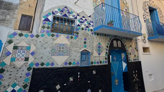 A house front in the Kasbah district of Mazara del Vallo, Sicily, covered in hand-set ceramic mosaic — coloured diamonds, patterned maiolica tiles and shells — with a Moorish-arched blue door and blue wrought-iron balconies above.