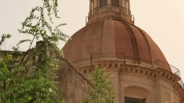 The large Baroque dome and lantern of the Monastero dei Benedettini church of San Nicolò l'Arena in Catania, Sicily, seen from below through green foliage.
