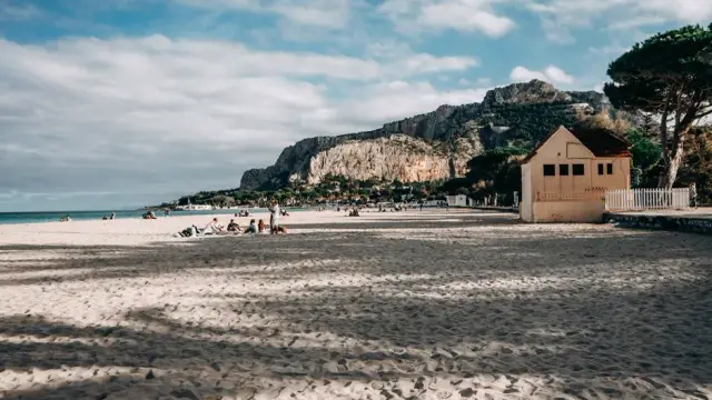 Mondello beach near Palermo, Sicily, with its sandy shore, a liberty-style bathhouse pavilion, and Monte Pellegrino rising behind it under a partly cloudy sky.