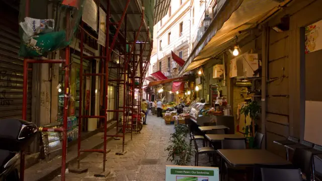 A narrow lane in Palermo's Vucciria market lined with food stalls, string lights, striped awnings, and outdoor restaurant tables set up for the evening.