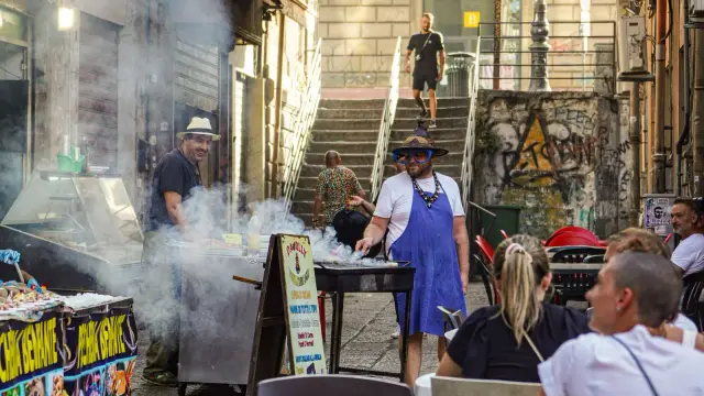 A street food stall in Palermo with barbecue smoke rising from the grill, handwritten signs advertising stigghiola and pani ca meusa, and people waiting on the steps of the alley behind.
