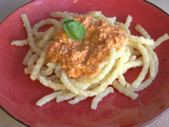 Busiate with Pesto alla Trapanese on a red plate, garnished with a basil leaf — the coarse, reddish tomato-and-almond sauce sits visibly chunky on top of the pasta.
