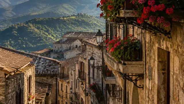 A narrow stone street in Petralia Soprana lined with flower-covered wrought-iron balconies, with the layered peaks of the Madonie mountains in the background.