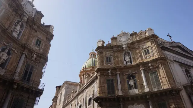 Two curved Baroque facades of Quattro Canti in Palermo meeting at a street corner, decorated with statues in niches and a green dome visible behind, under a clear blue sky.