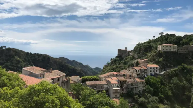 A hilltop Sicilian village with terracotta rooftops spilling down a green ridge, a stone castle tower perched on the crest above it, and the sea visible in the distance.