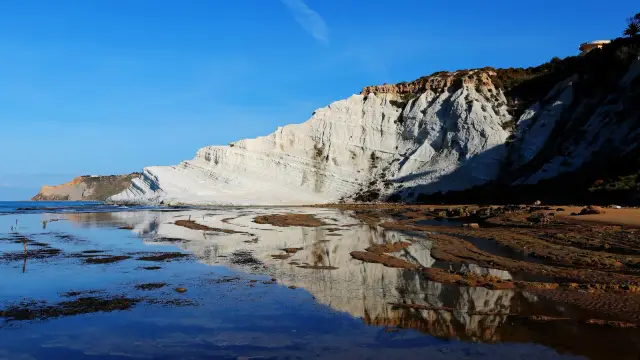 The white marl cliff of Scala dei Turchi near Realmonte, Sicily, its stepped terraces glowing in low sunlight and mirrored in a shallow pool of water on the rocky foreshore, under a deep blue sky.