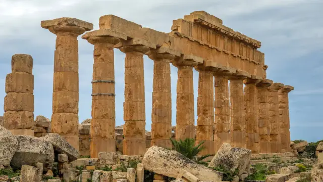 Weathered Doric columns and a partial entablature of Temple C on the acropolis of Selinunte, Sicily, standing among fallen stone blocks under a pale blue sky.