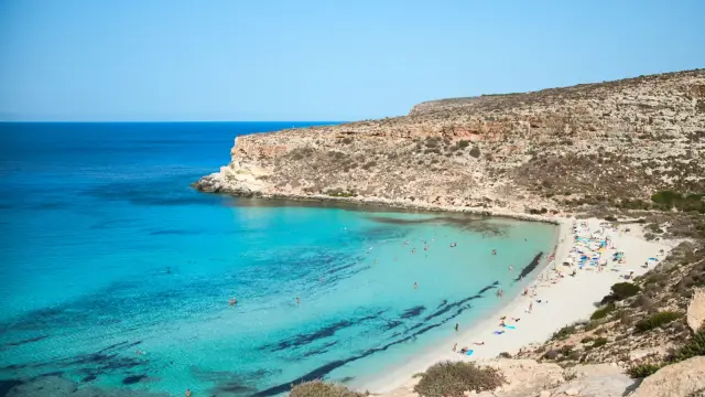 Aerial view of Spiaggia dei Conigli on Lampedusa, showing a crescent of white sand and exceptionally clear turquoise-to-deep-blue water surrounded by pale rocky cliffs.