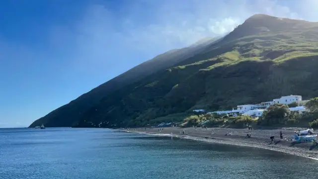 View from the sea toward Stromboli: a green volcanic slope drops steeply to the water, a small white village sits on the shore, with clear turquoise water in front.