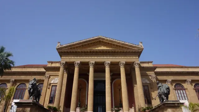 The grand neoclassical facade of Teatro Massimo in Palermo, with its wide front steps, Corinthian columns and bronze lion statues under a clear blue sky.