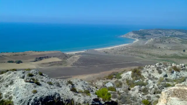 An elevated view of the wild, undeveloped coastline of the Torre Salsa nature reserve near Siculiana, Sicily, showing clay cliffs, an empty sandy beach, and turquoise water curving toward a distant headland.