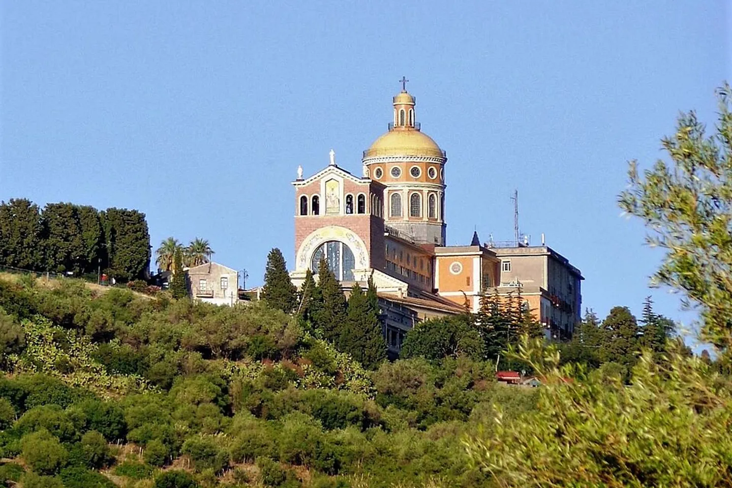 The sanctuary of the Black Madonna at Tindari in Sicily, its golden dome rising above a wooded hillside against a clear blue sky.