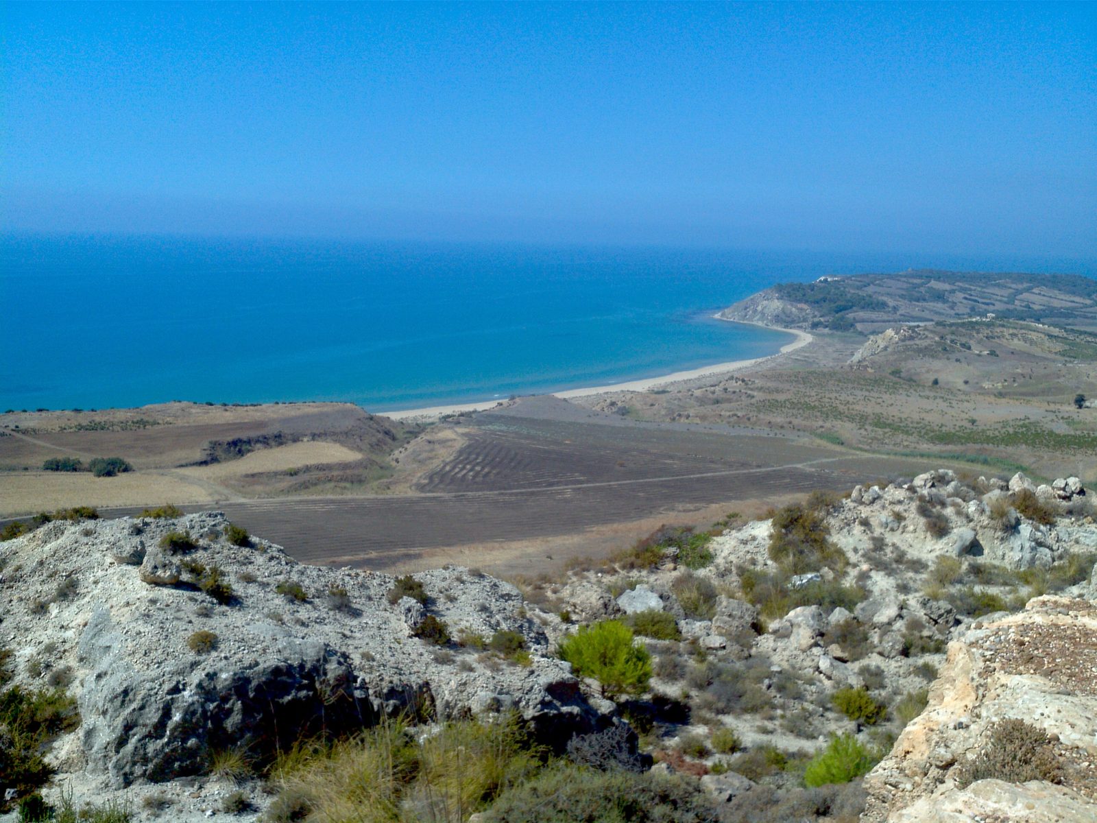 An elevated view of the wild, undeveloped coastline of the Torre Salsa nature reserve near Siculiana, Sicily, showing clay cliffs, an empty sandy beach, and turquoise water curving toward a distant headland.