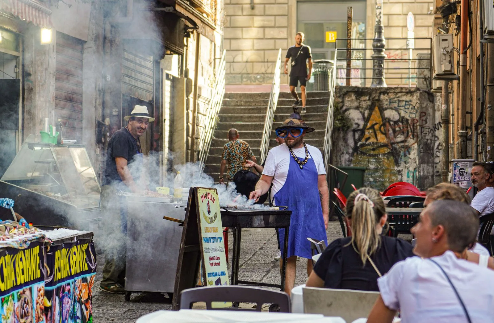 Street food grill with smoke rising over a lively alley in Palermo, Sicily, with people sitting at nearby tables.